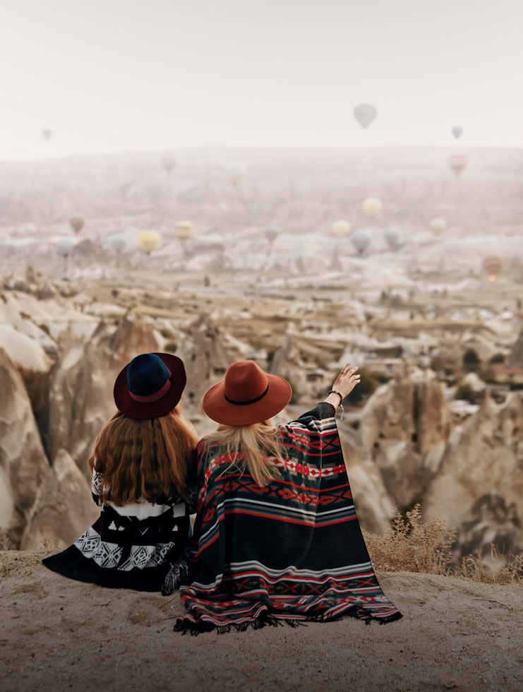 women-in-hats-sitting-on-a-hill-enjoying-flying-hot-air-balloons-view-at-cappadocia-infocappadocia.com